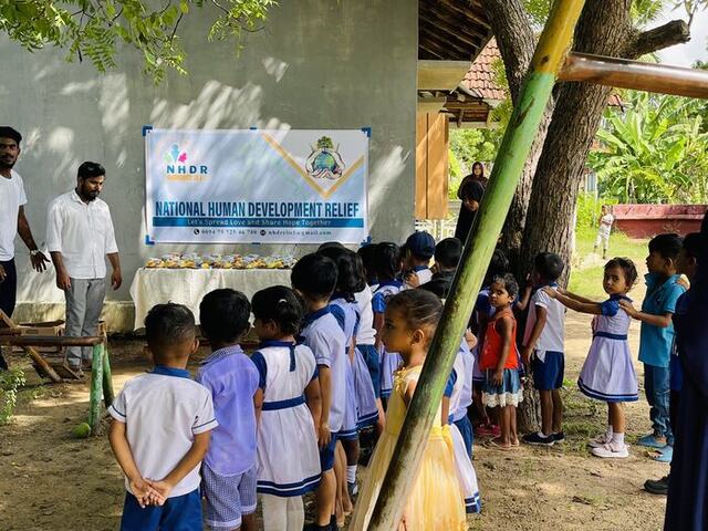 Schoolchildren lined up under a tree beside an NHDR banner