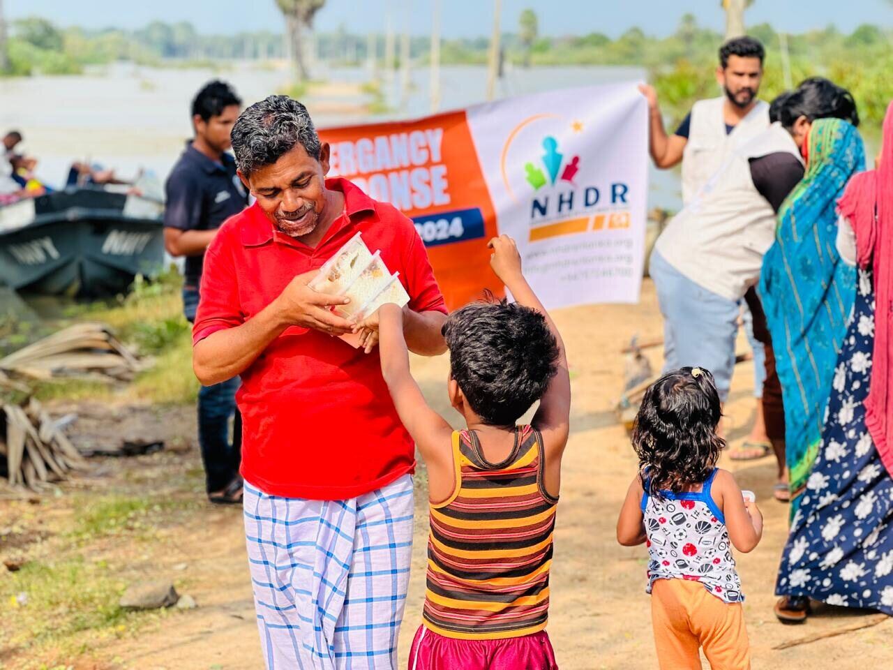 NHDR volunteers handing out meal boxes to children beside floodwaters, December 2024